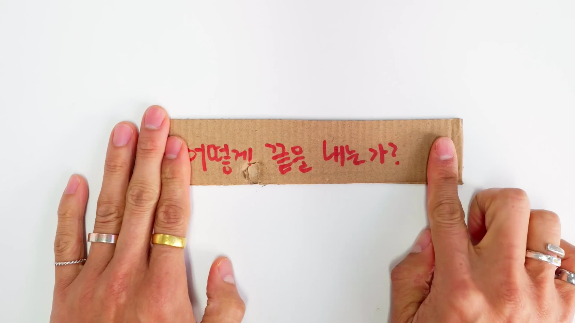 흰색 배경 위에서 양손이 골판지 조각의 양끝을 잡고 있으며, 골판지에는 빨간색 펜으로 '어떻게 끝을 내는가?'라는 문구가 적혀 있다.