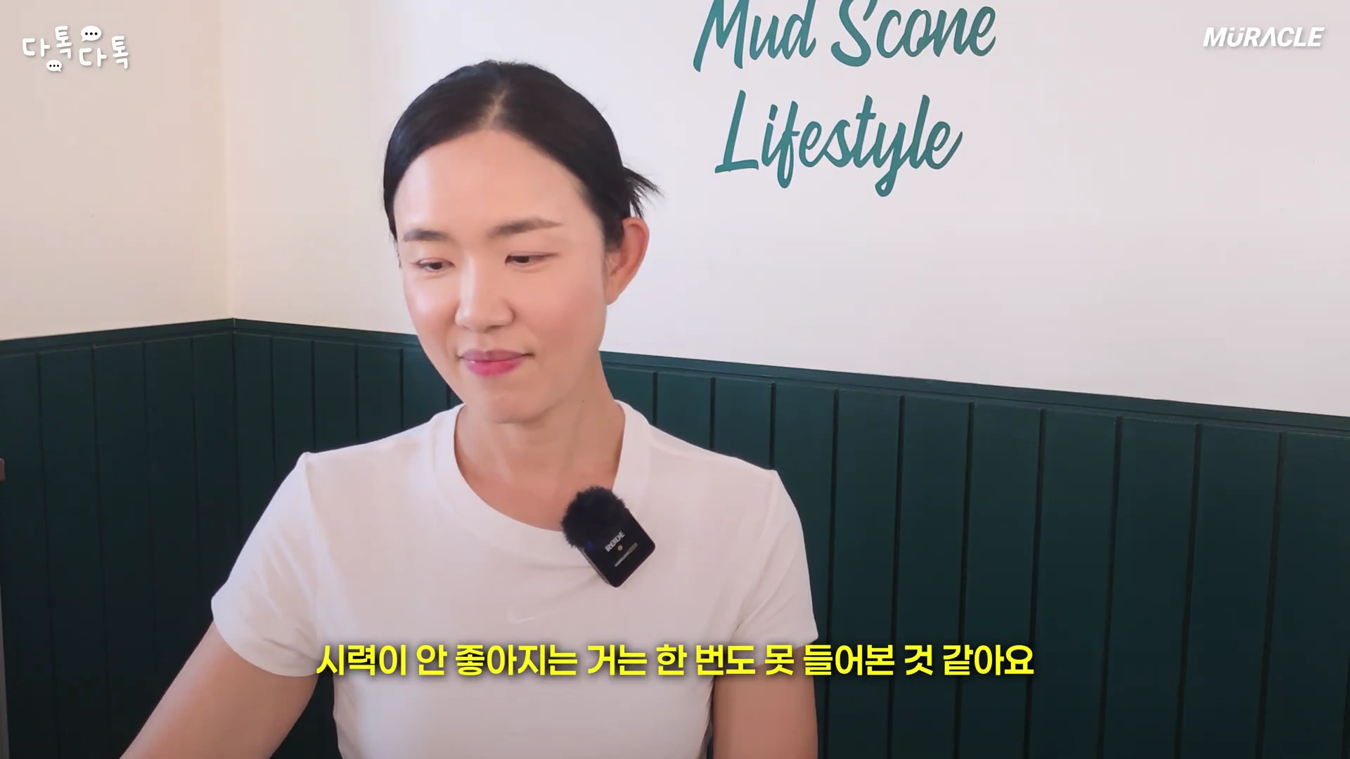 흰색 티셔츠를 입고 핀 마이크를 착용한 여성이 실내에서 이야기를 나누고 있으며, 하단에는 노란색 한글 자막이 표시되어 있습니다.