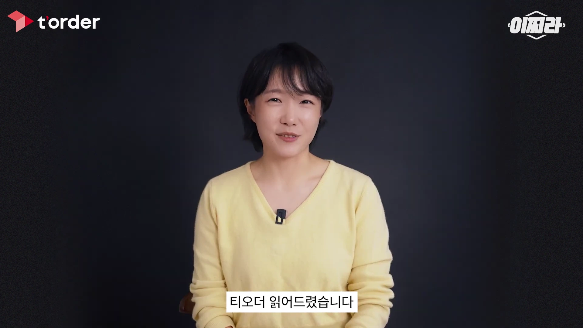 검은색 배경 앞에서 노란색 니트를 입은 단발머리 여성이 카메라를 정면으로 응시하며 이야기하고 있습니다.