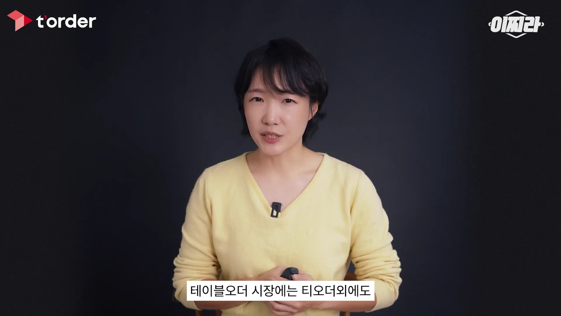 노란색 니트를 입은 단발머리 여성이 검은색 배경 앞에서 정면을 바라보며 이야기하고 있다.