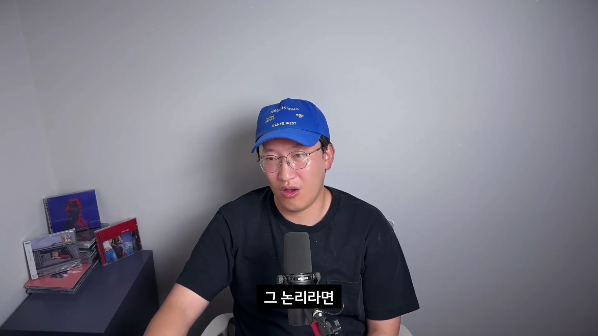 파란색 야구 모자와 안경을 착용한 남성이 마이크 앞에 앉아 이야기를 하고 있으며, 왼쪽 옆 탁자 위에는 여러 개의 음반 케이스가 놓여 있다.