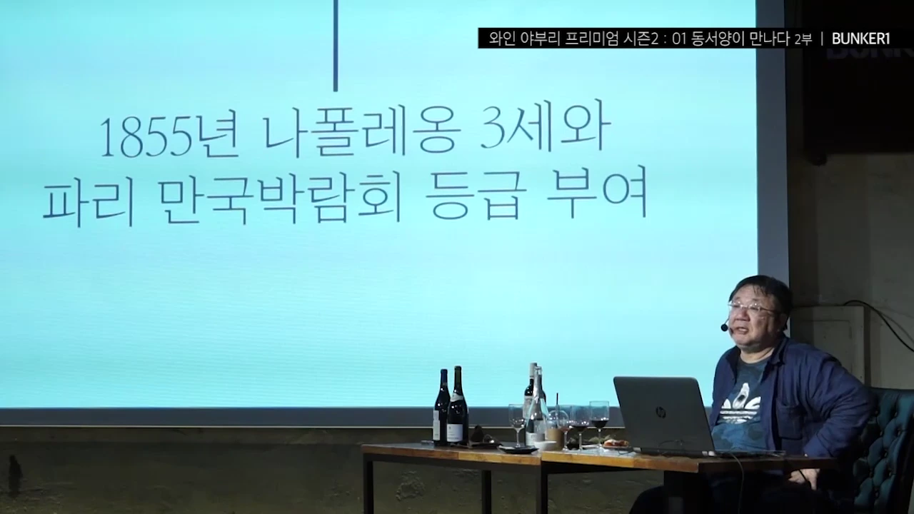 강단에 앉은 중년 남성이 1855년 나폴레옹 3세와 파리 만국박람회 등급 부여라는 글자가 적힌 대형 스크린을 등지고 앉아 있는 모습.