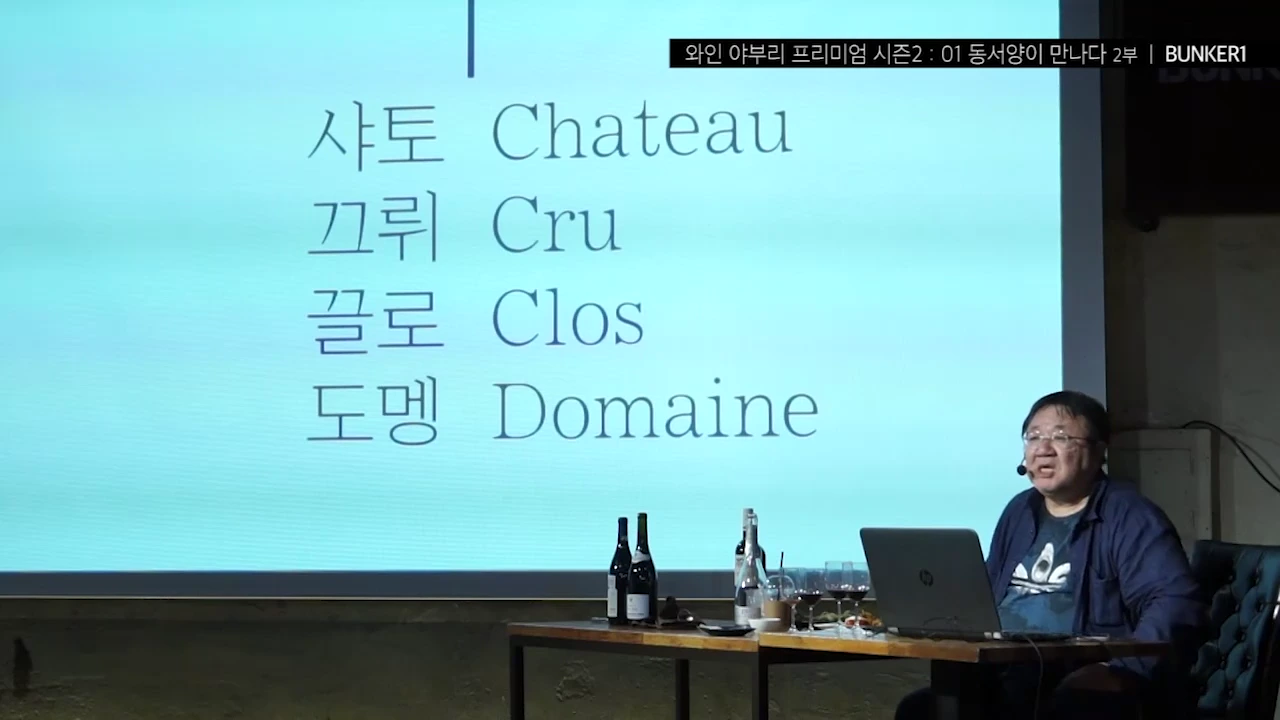 강연자가 노트북 앞에 앉아 있고, 뒤편 대형 화면에는 '샤토(Chateau)', '끄뤼(Cru)', '끌로(Clos)', '도멘(Domaine)'이라는 단어가 한국어와 영어로 나란히 적혀 있습니다.