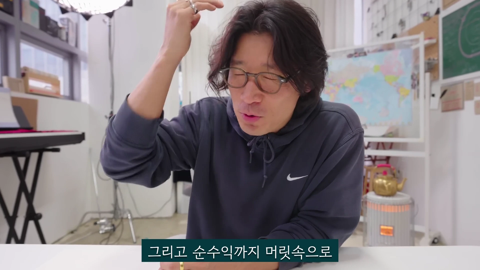 안경을 쓴 남성이 후드티를 입고 앉아 손을 들어 머리 근처에서 제스처를 취하며 이야기하는 모습의 영상 캡처 화면입니다.