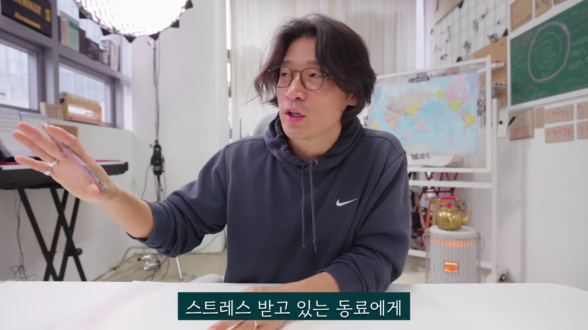 안경을 쓴 남성이 밝은 사무실 책상에 앉아 손짓하며 이야기를 나누고 있는 모습이 담긴 영상 캡처 화면입니다. 화면 하단에는 '스트레스 받고 있는 동료에게'라는 자막이 있습니다.