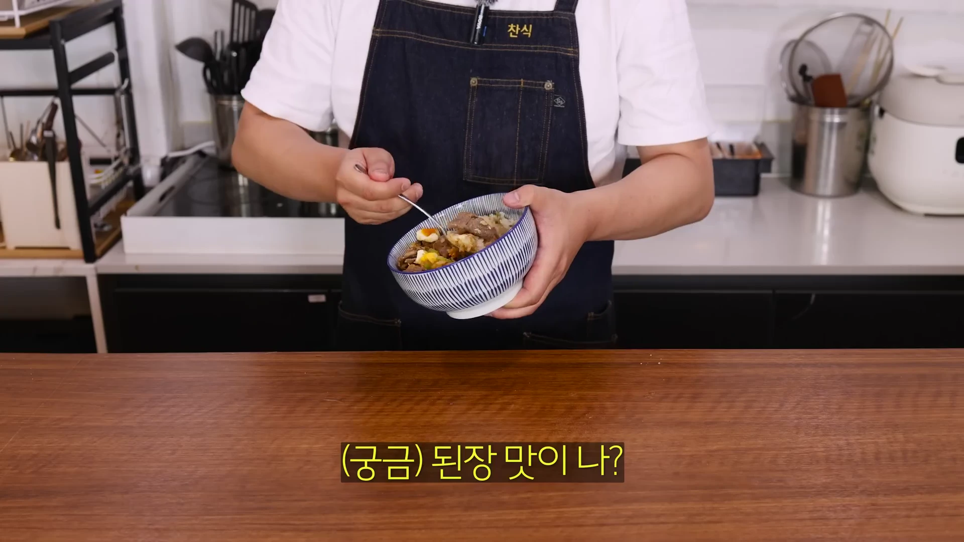 앞치마를 두른 요리사가 주방에서 미소돼지고기 덮밥이 담긴 줄무늬 사발을 들고 숟가락으로 재료를 섞고 있다.