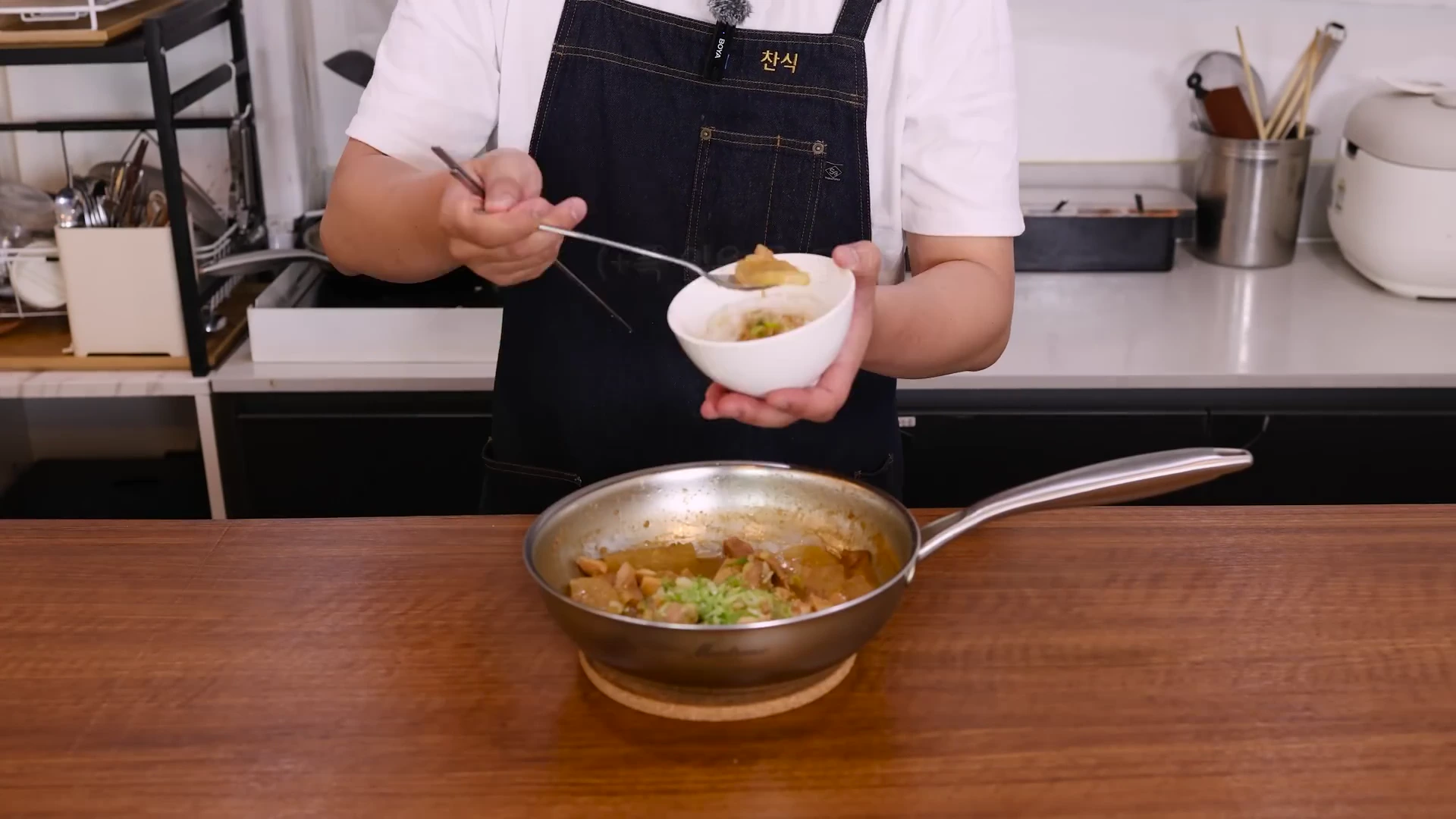 앞치마를 두른 요리사가 숟가락으로 프라이팬에 담긴 미소 닭고기 조림을 떠서 흰색 그릇에 옮겨 담고 있는 모습.