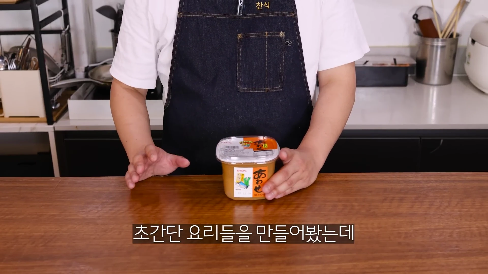 앞치마를 두른 요리사가 주방 조리대 위에서 일본식 된장인 미소 된장 용기를 양손으로 가리키고 있는 모습이다.