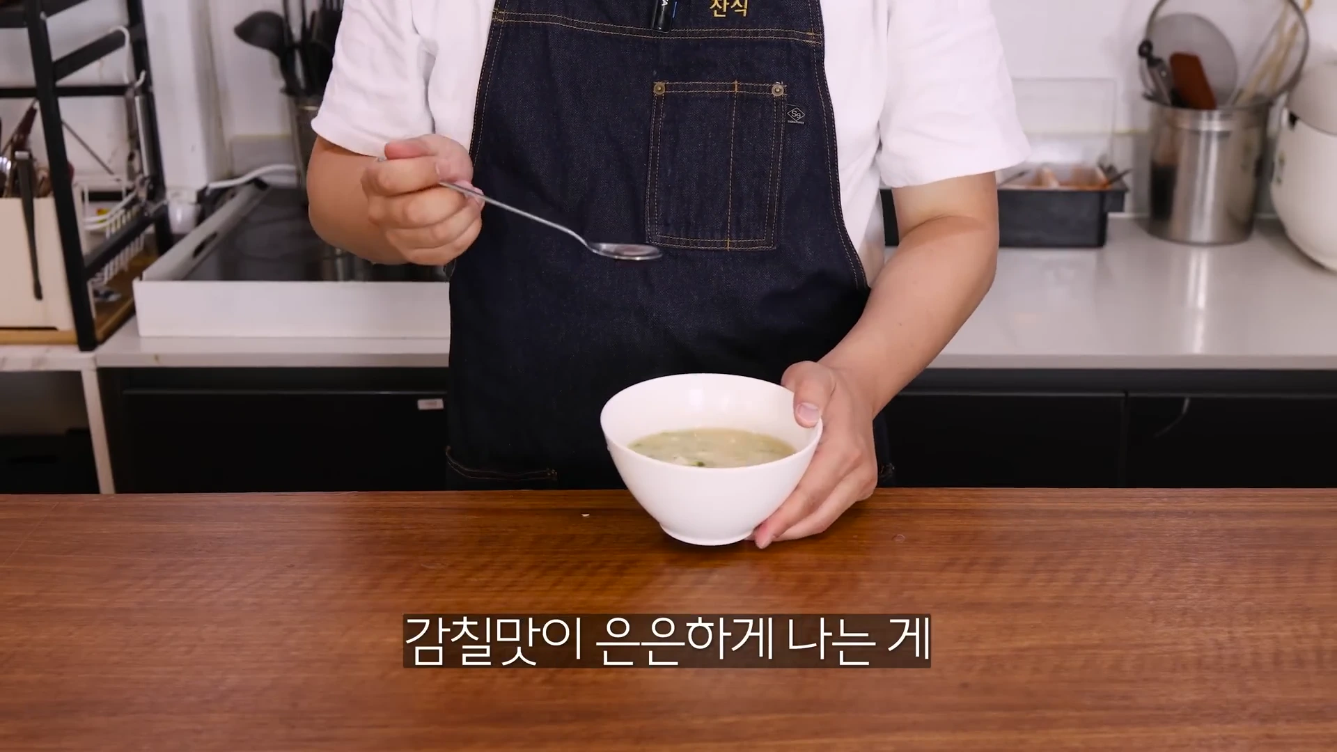 앞치마를 입은 사람이 미소 장국이 담긴 흰색 그릇을 들고 숟가락으로 국물을 뜨고 있는 모습