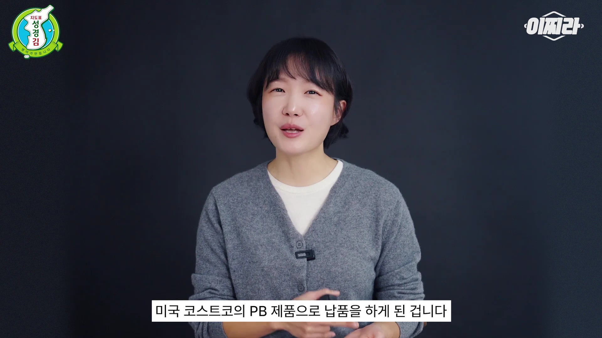 단발머리의 여성이 회색 브이넥 가디건을 입고 검은색 배경 앞에서 카메라를 보고 말하고 있으며, 하단에는 '미국 코스트코의 PB 제품으로 납품을 하게 된 겁니다'라는 자막이 떠 있습니다.