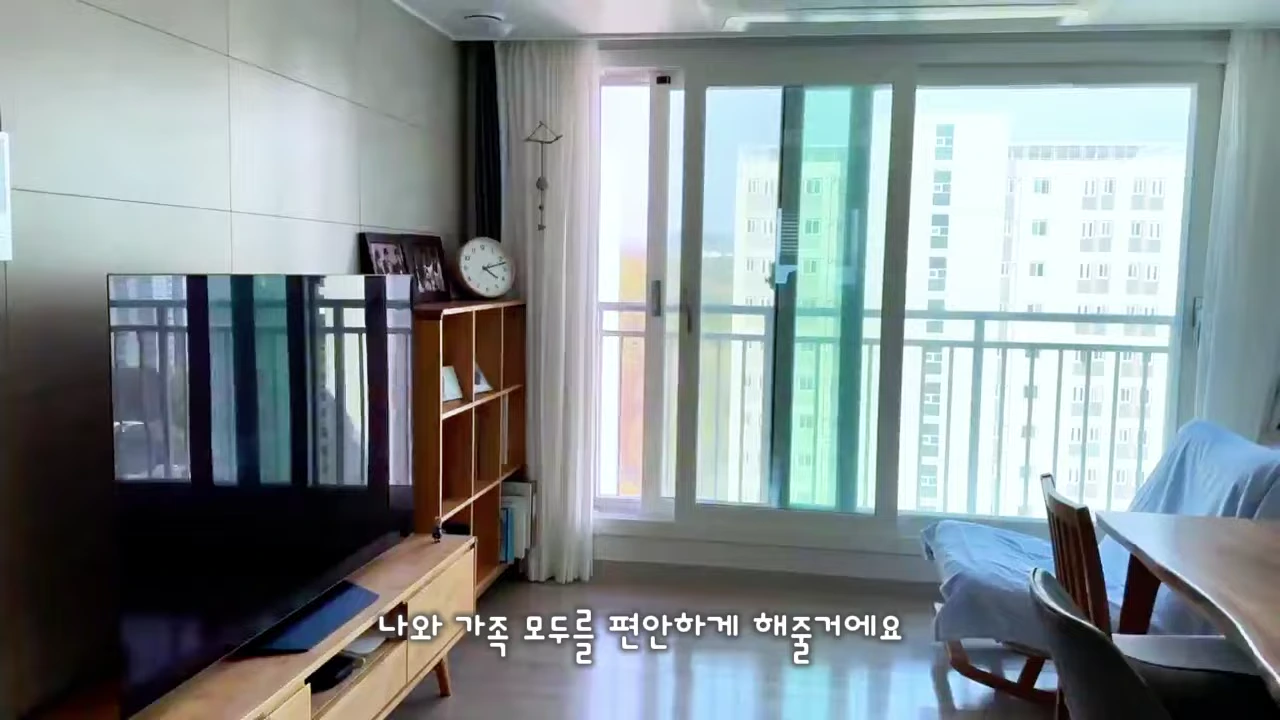 깔끔하게 정리된 거실에 텔레비전과 나무 선반, 창가에 놓인 의자가 배치된 현대적인 아파트 내부 모습
