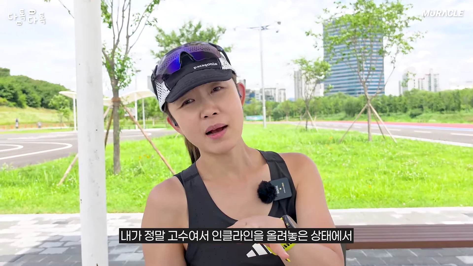 러닝 복장을 한 여성이 야외 공원을 배경으로 카메라를 보며 이야기를 나누고 있다.