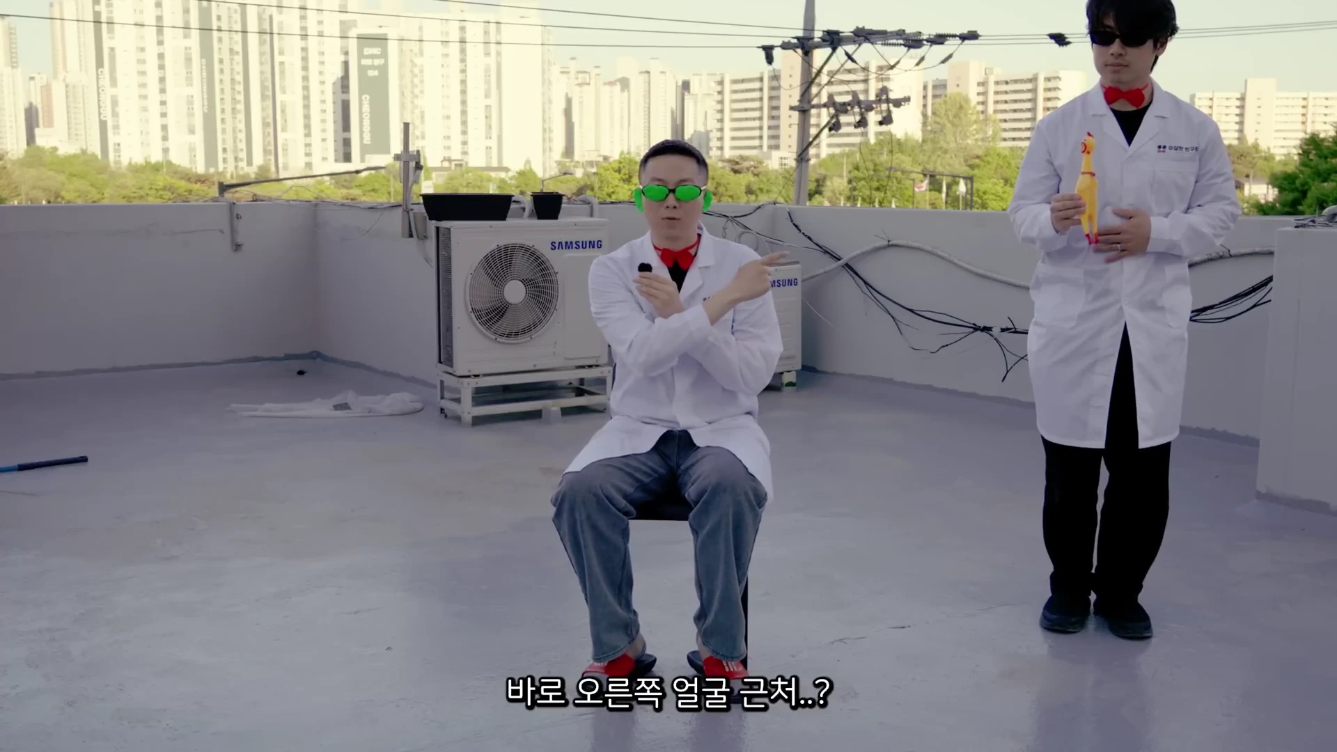 흰 가운을 입은 두 남자가 옥상에서 실험하는 모습이며, 한 남자는 초록색 안경을 쓰고 의자에 앉아 있고 다른 남자는 옆에서 장난감 닭을 들고 서 있습니다.