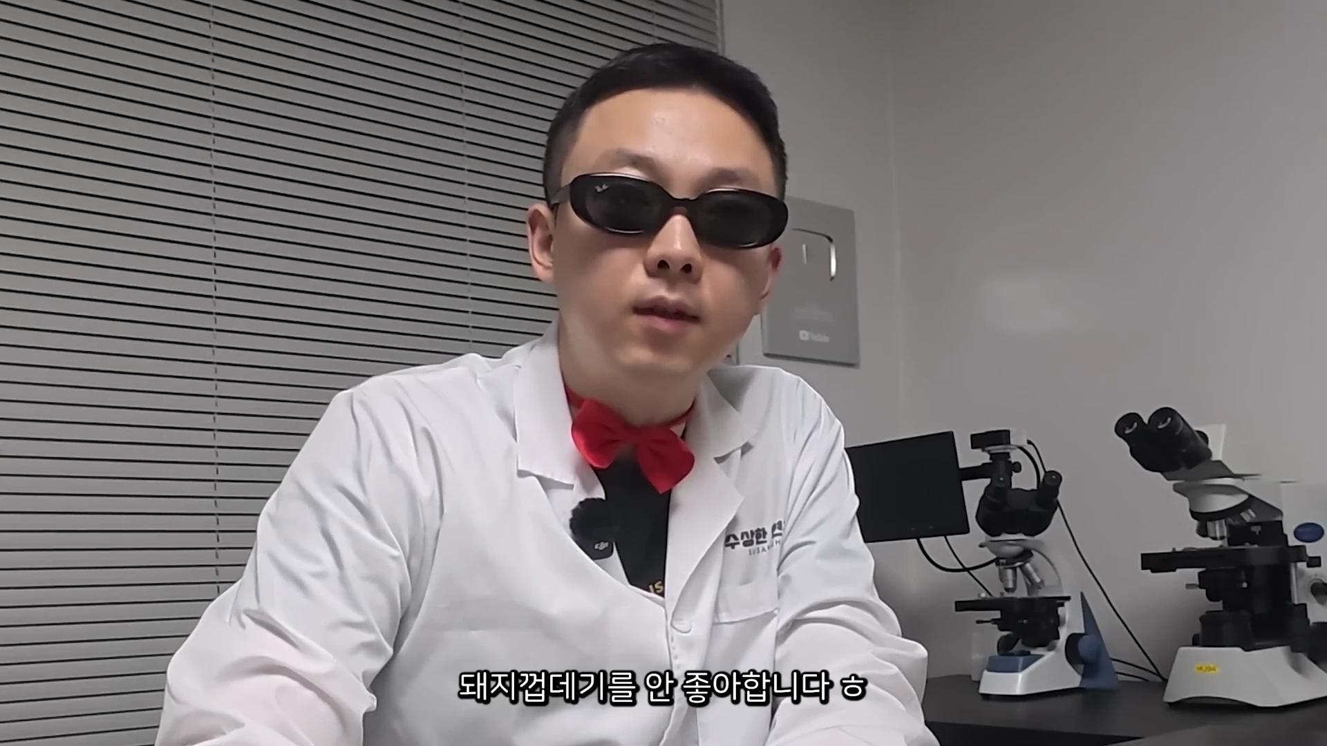 흰색 가운과 빨간색 나비넥타이를 착용한 남성이 선글라스를 쓰고 테이블 앞에 앉아 화면을 응시하고 있으며, 배경에는 블라인드와 현미경이 보인다.