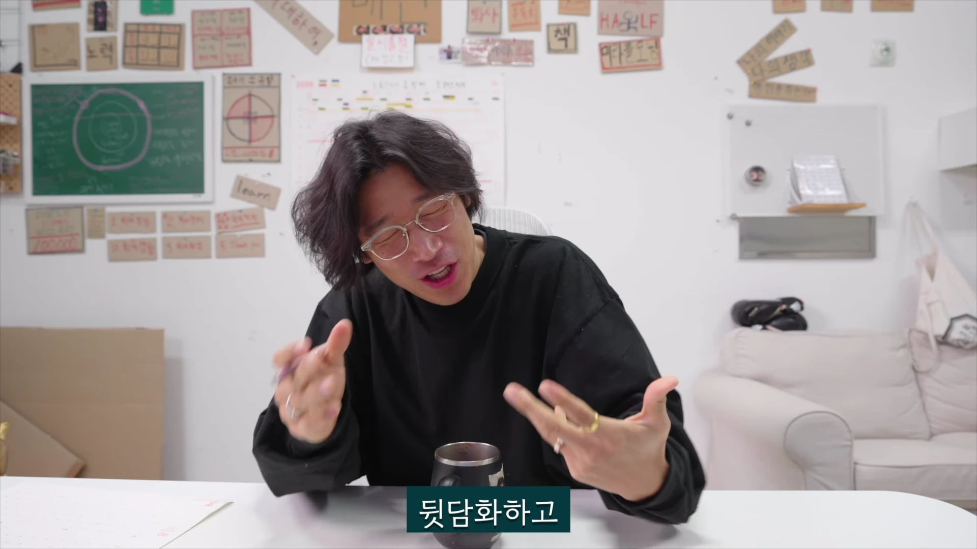 안경을 쓴 남성이 책상 앞에 앉아 손짓하며 이야기를 나누고 있으며, 배경의 벽에는 다양한 메모지와 게시물이 붙어 있고 하단에는 '뒷담화하고'라는 자막이 보입니다.