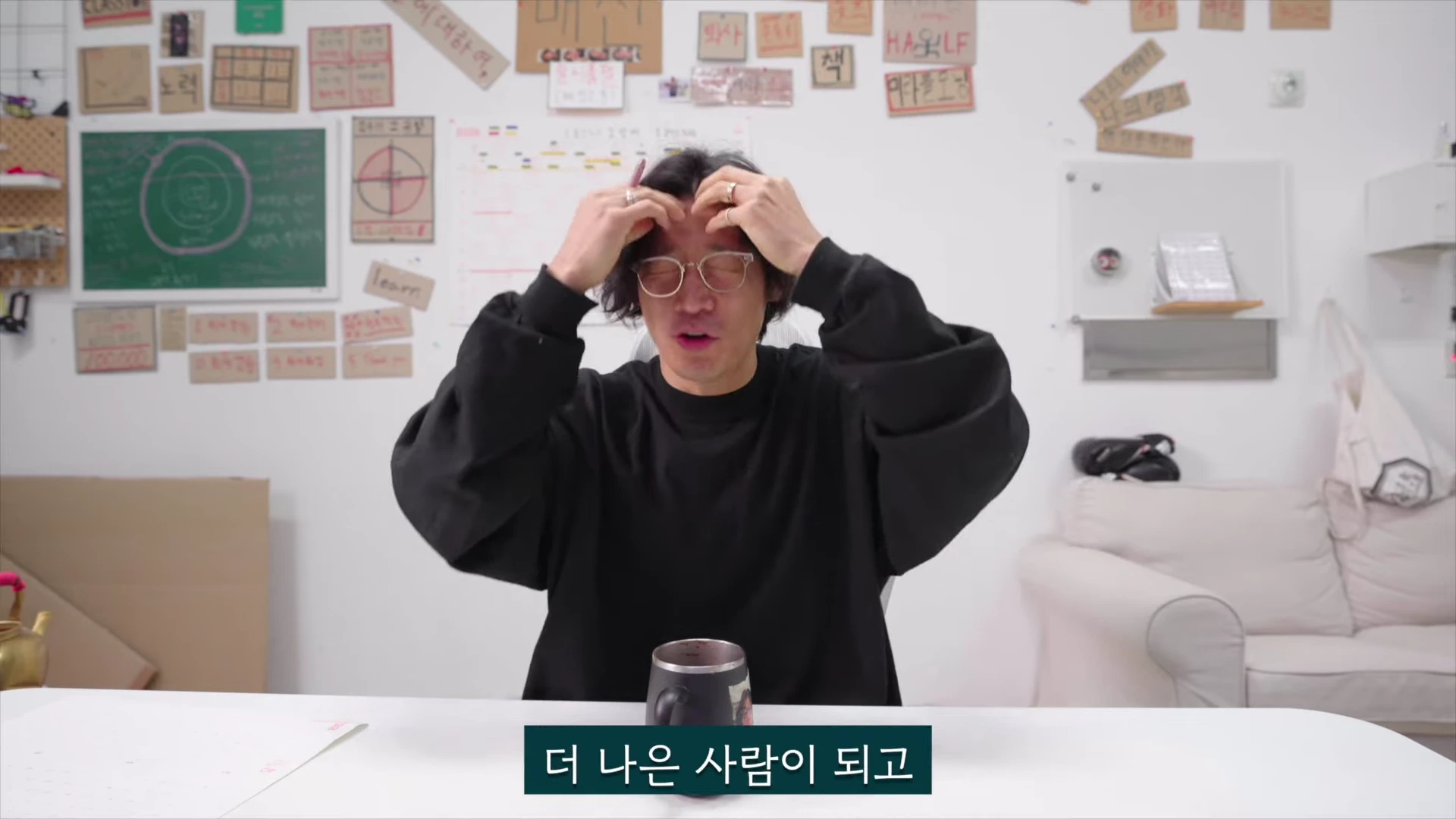 흰색 배경의 작업실에서 검은색 긴팔 티셔츠를 입고 안경을 쓴 남성이 양손으로 머리카락을 쓸어 올리며 고민하는 듯한 표정을 짓고 있다.