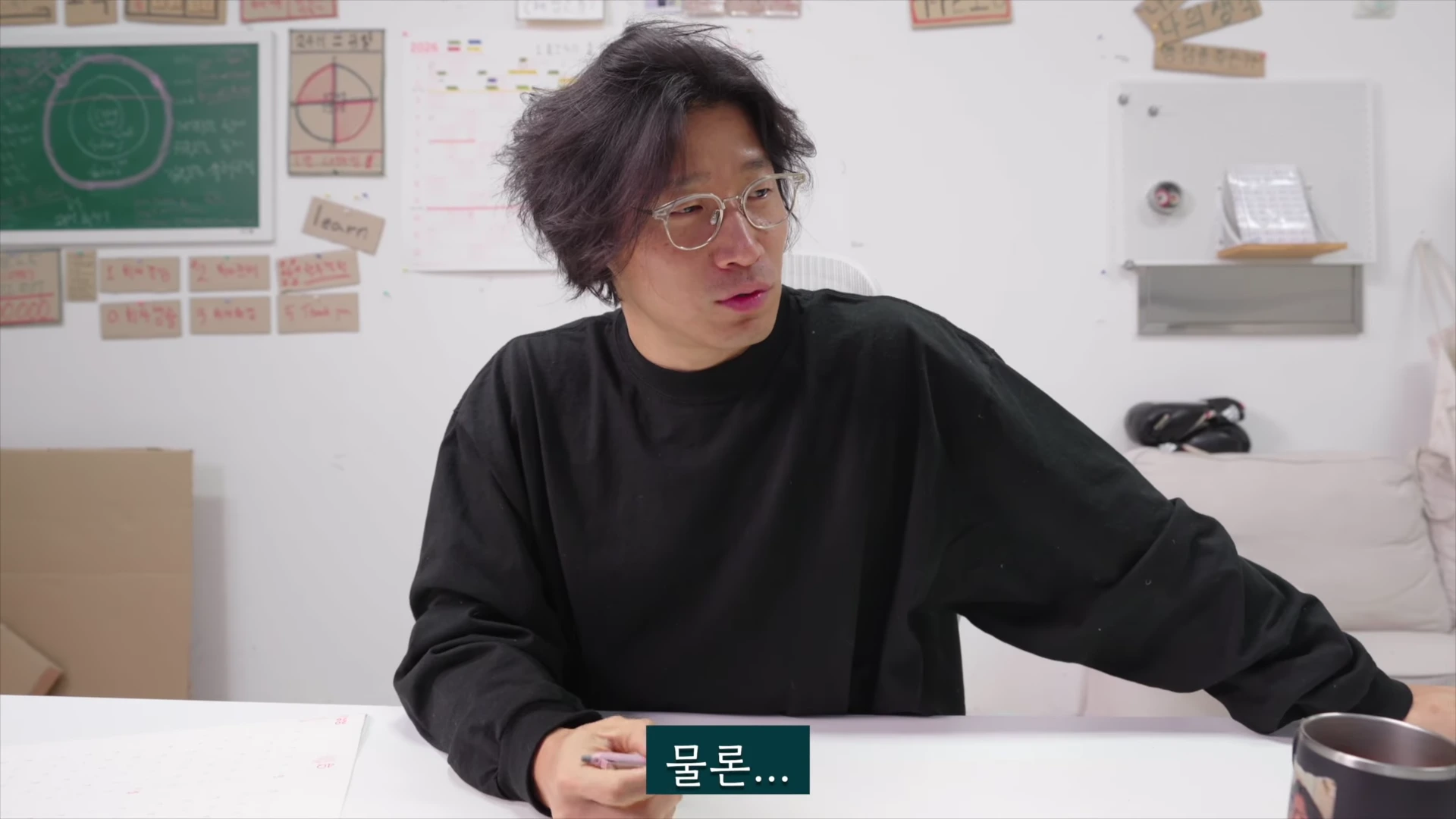 검은색 긴팔 티셔츠를 입고 안경을 쓴 남성이 책상에 앉아 옆을 바라보며 이야기하고 있으며, 뒤쪽 벽면에는 학습 자료와 메모가 붙어 있다.