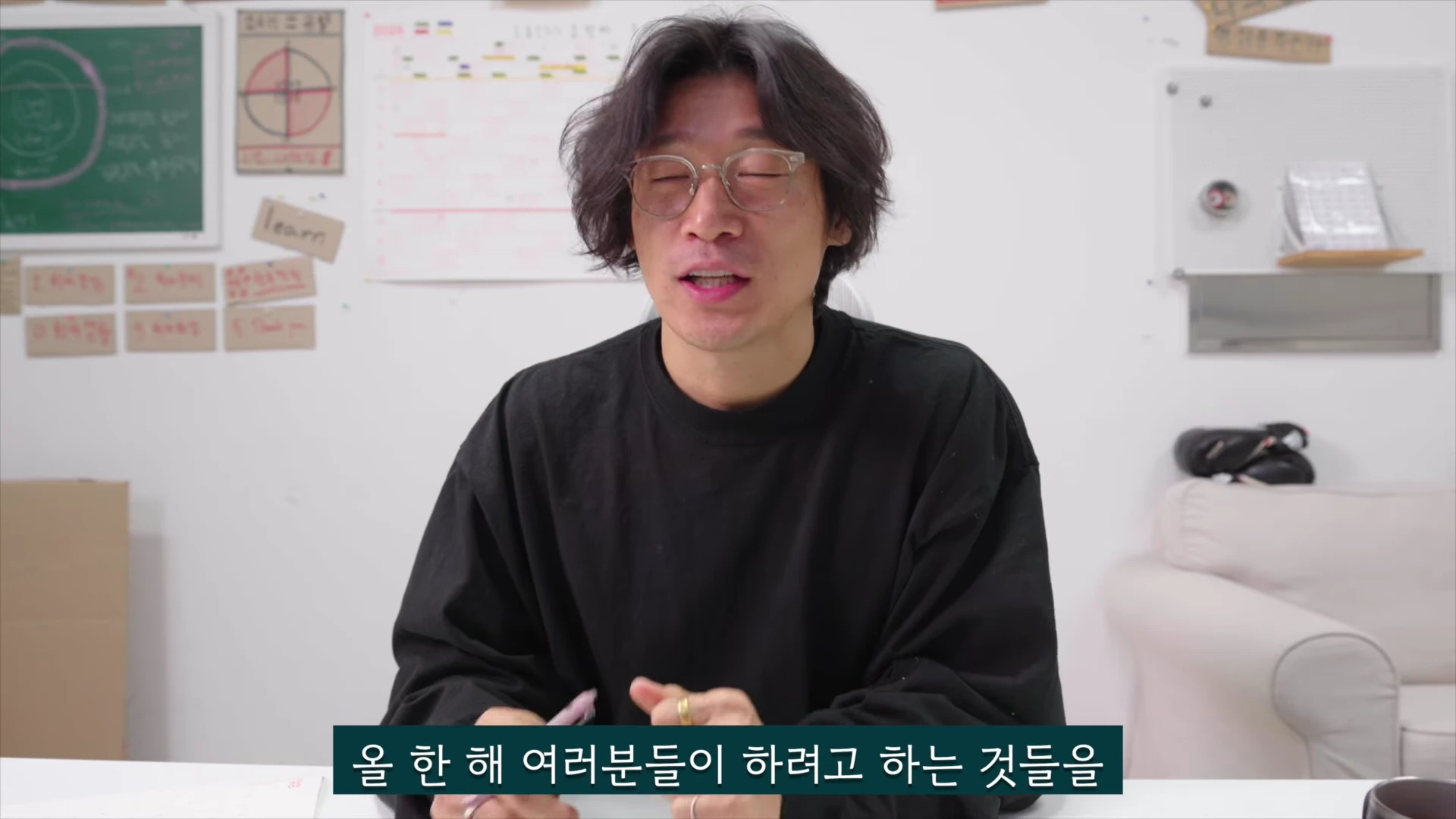 안경을 쓴 남성이 흰 벽을 배경으로 앉아 차분하게 이야기를 하고 있으며, 뒤쪽 벽면에는 메모지와 학습 목표 등이 적힌 게시물이 붙어 있다.