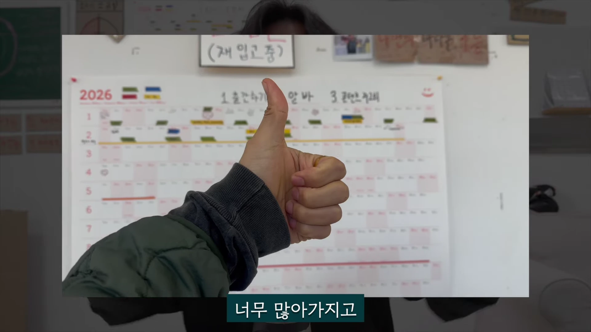 한 사람의 손이 엄지손가락을 치켜세우고 있으며 그 뒤로 2026년 계획이 적힌 달력과 메모가 붙은 벽이 보인다.