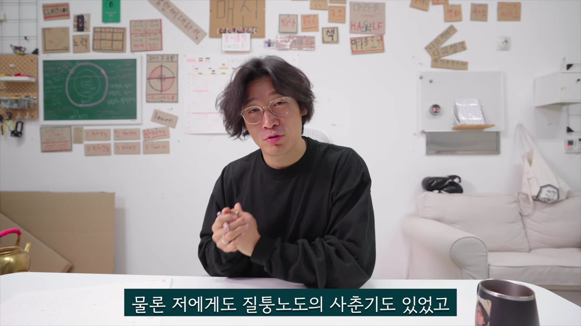 안경을 쓴 남성이 흰색 배경을 배경으로 앉아 손을 모으고 이야기를 나누는 모습이며, 벽에는 다양한 문구가 적힌 메모지들이 붙어 있습니다.