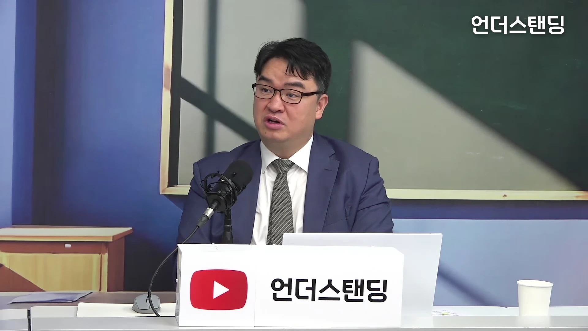 안경을 쓴 남성이 스튜디오에 앉아 마이크 앞에서 이야기하고 있으며 앞에는 '언더스탠딩' 로고가 적힌 패널이 놓여 있습니다.