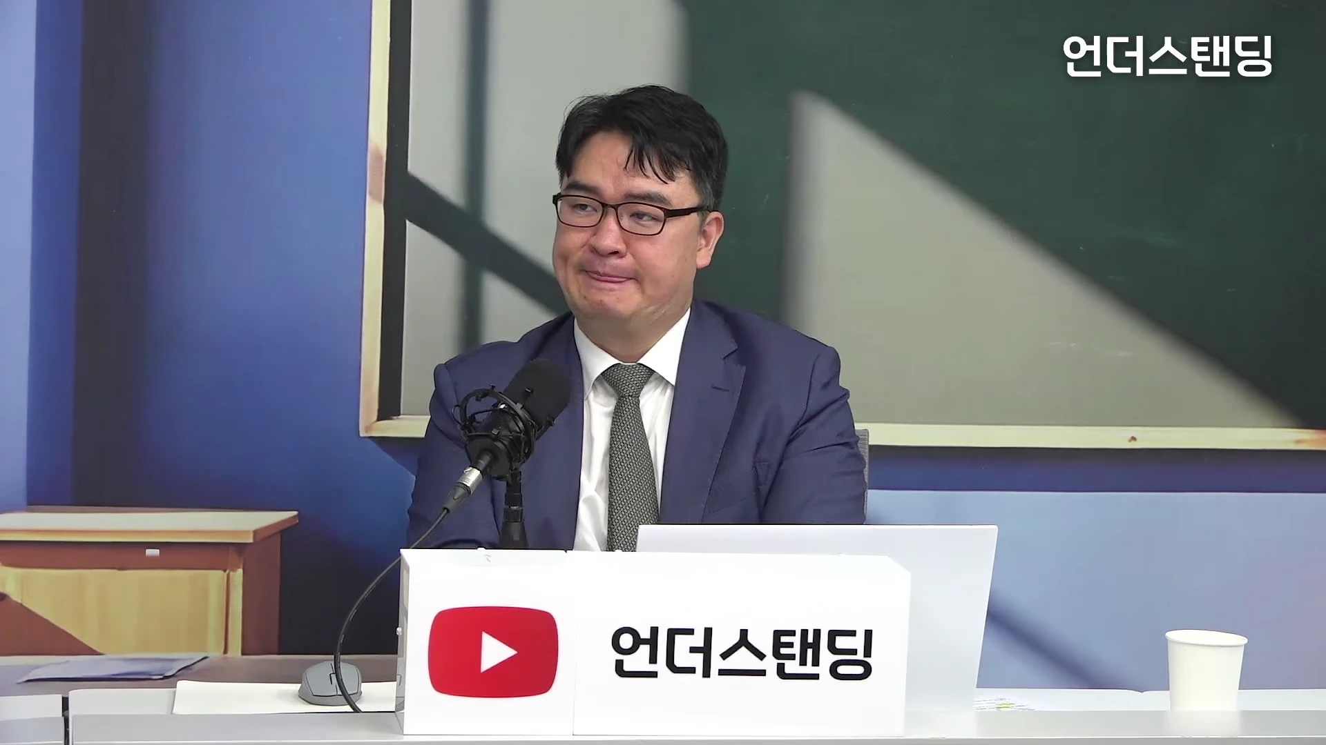 안경을 쓴 남성이 스튜디오에 앉아 마이크 앞에 대기하고 있는 모습