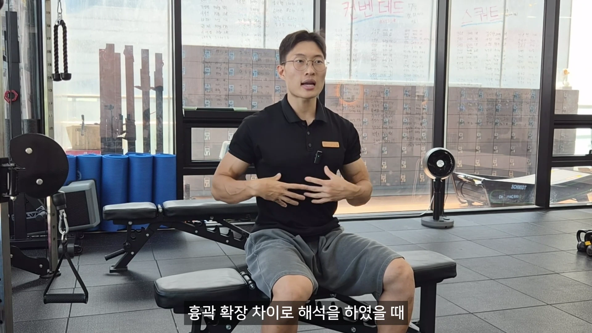 운동복 차림의 남성이 헬스장에서 앉아 자신의 가슴과 흉곽 부위를 손으로 가리키며 설명하는 모습