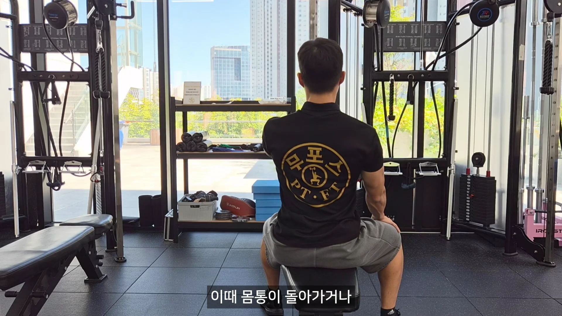 등 뒤에서 본 헬스장에서 검은색 상의를 입은 남성이 원암 시티드 로우 기구 앞에 앉아 손잡이를 당기는 동작을 보여주는 영상 프레임이다.