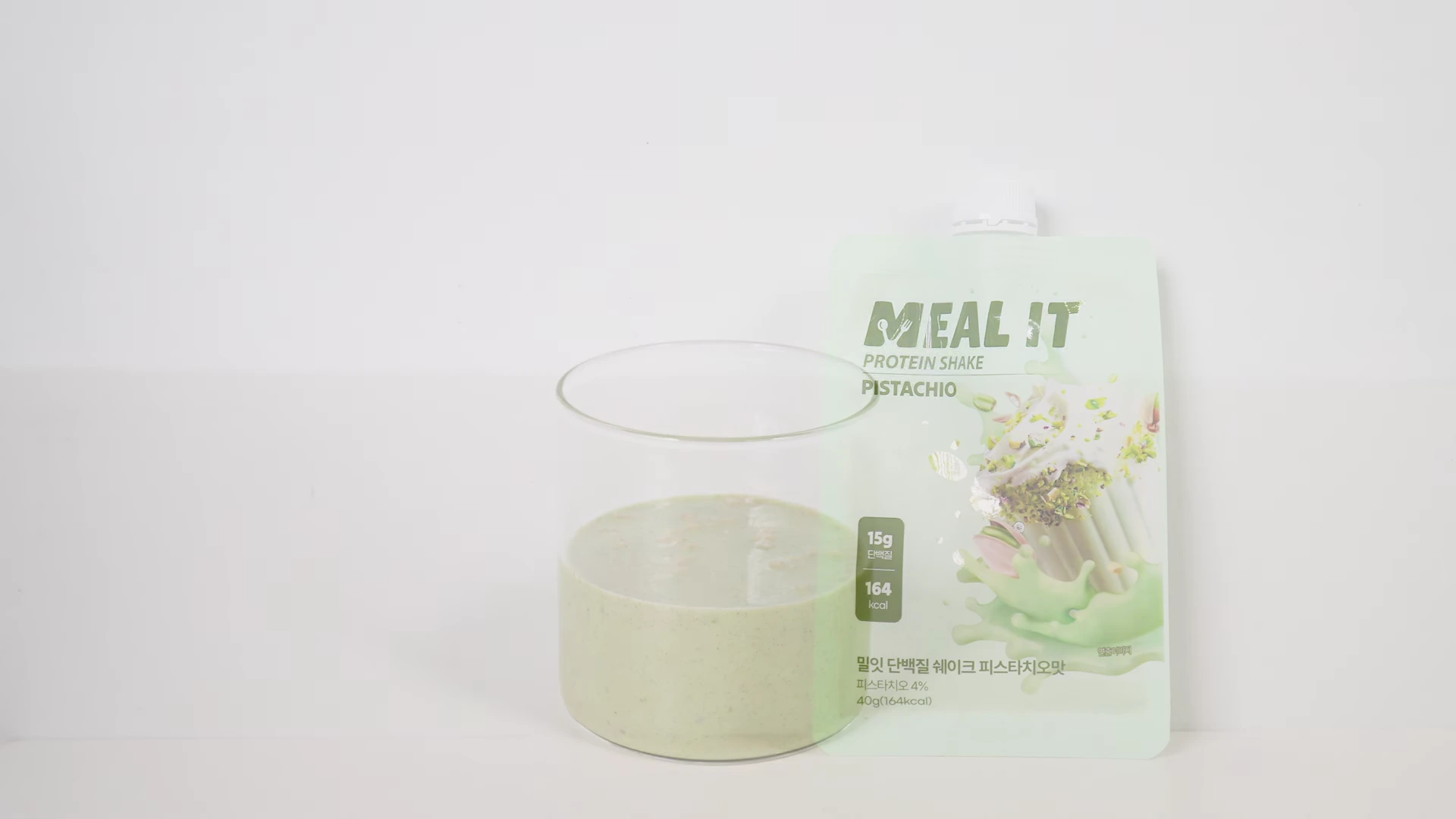 연한 녹색의 밀릿 단백질 쉐이크 피스타치오맛 파우치 제품과 그 옆에 같은 색상의 내용물이 담긴 유리컵이 놓여 있는 모습입니다.