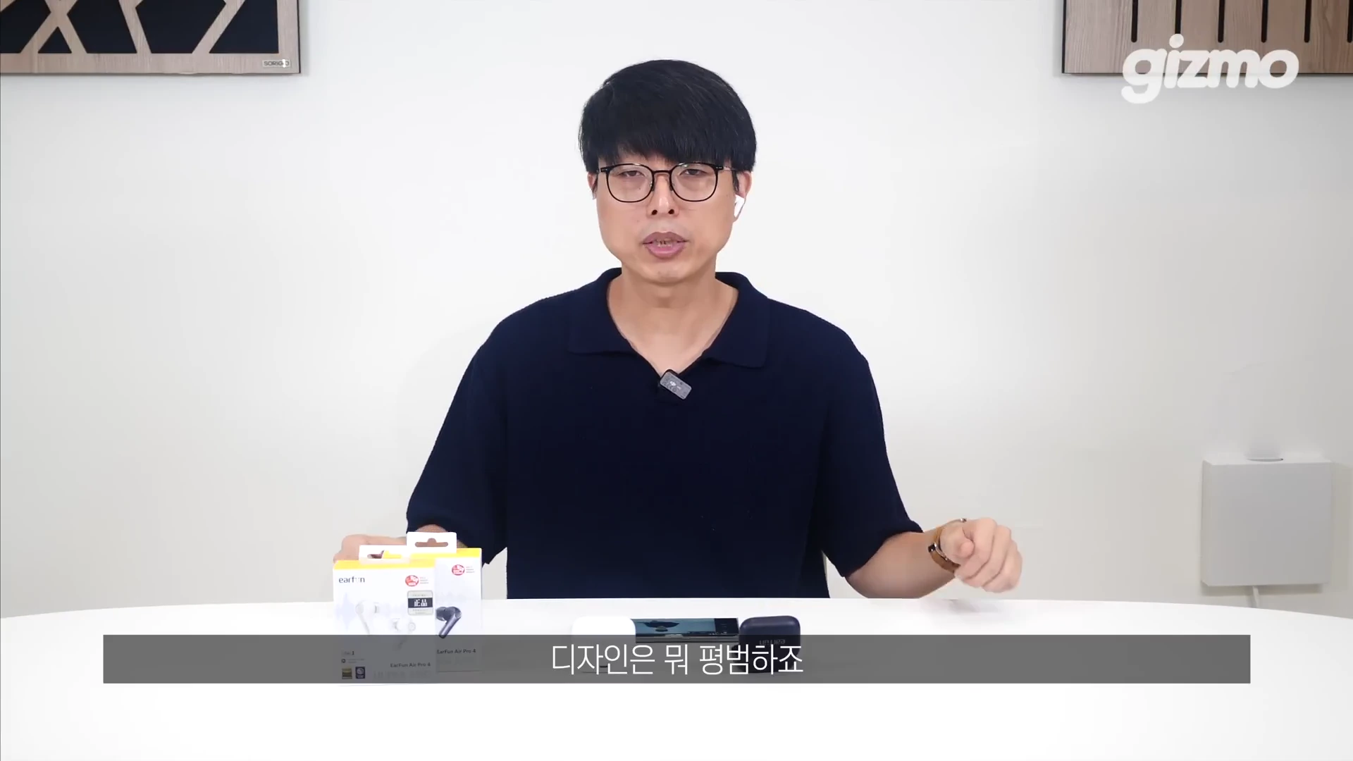 흰색 배경의 스튜디오에서 안경을 쓴 남성이 책상 앞에 앉아 있으며, 책상 위에는 무선 이어폰 패키지 박스와 충전 케이스가 놓여 있습니다.