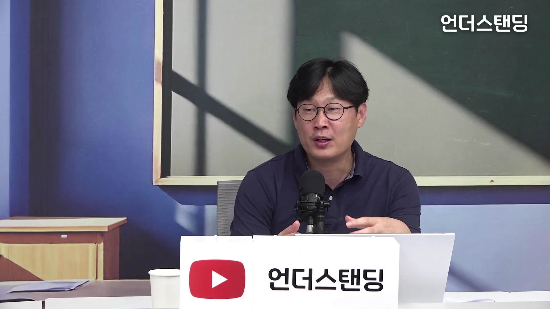 안경을 쓴 남성이 마이크 앞에 앉아 이야기를 나누고 있는 모습으로, 앞쪽에는 유튜브 로고와 채널명이 적힌 흰색 명패가 놓여 있다.