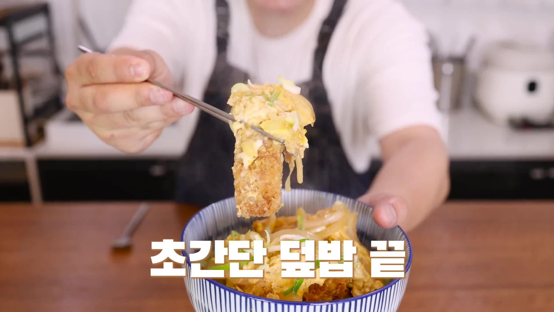 흰색 티셔츠와 앞치마를 입은 사람이 젓가락으로 카츠동의 돈가스 조각을 집어 카메라 앞으로 보여주고 있으며 화면 하단에는 흰색 자막이 적혀 있다.