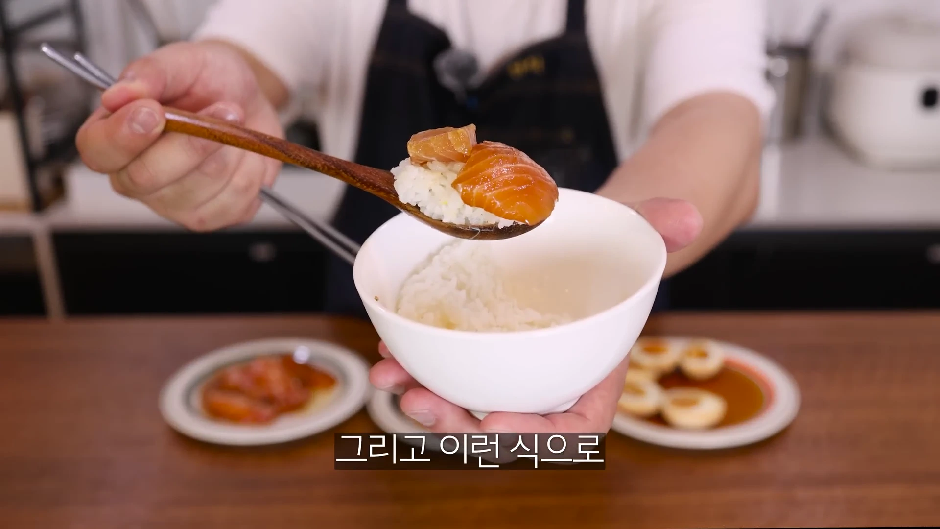 흰 쌀밥 위에 간장 절임 연어 한 점이 올려진 나무 숟가락을 한 손으로 들고 있는 모습. 배경에는 계란장과 다른 절임 요리가 담긴 접시들이 놓여 있다.