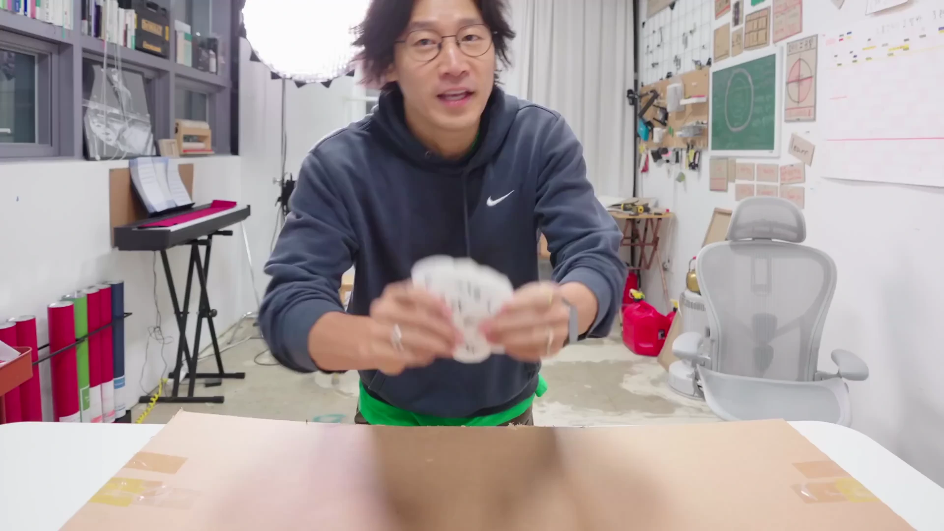 안경을 쓴 남성이 스튜디오 안에서 카메라를 향해 무언가를 설명하며 양손으로 작은 종이를 들고 있습니다.