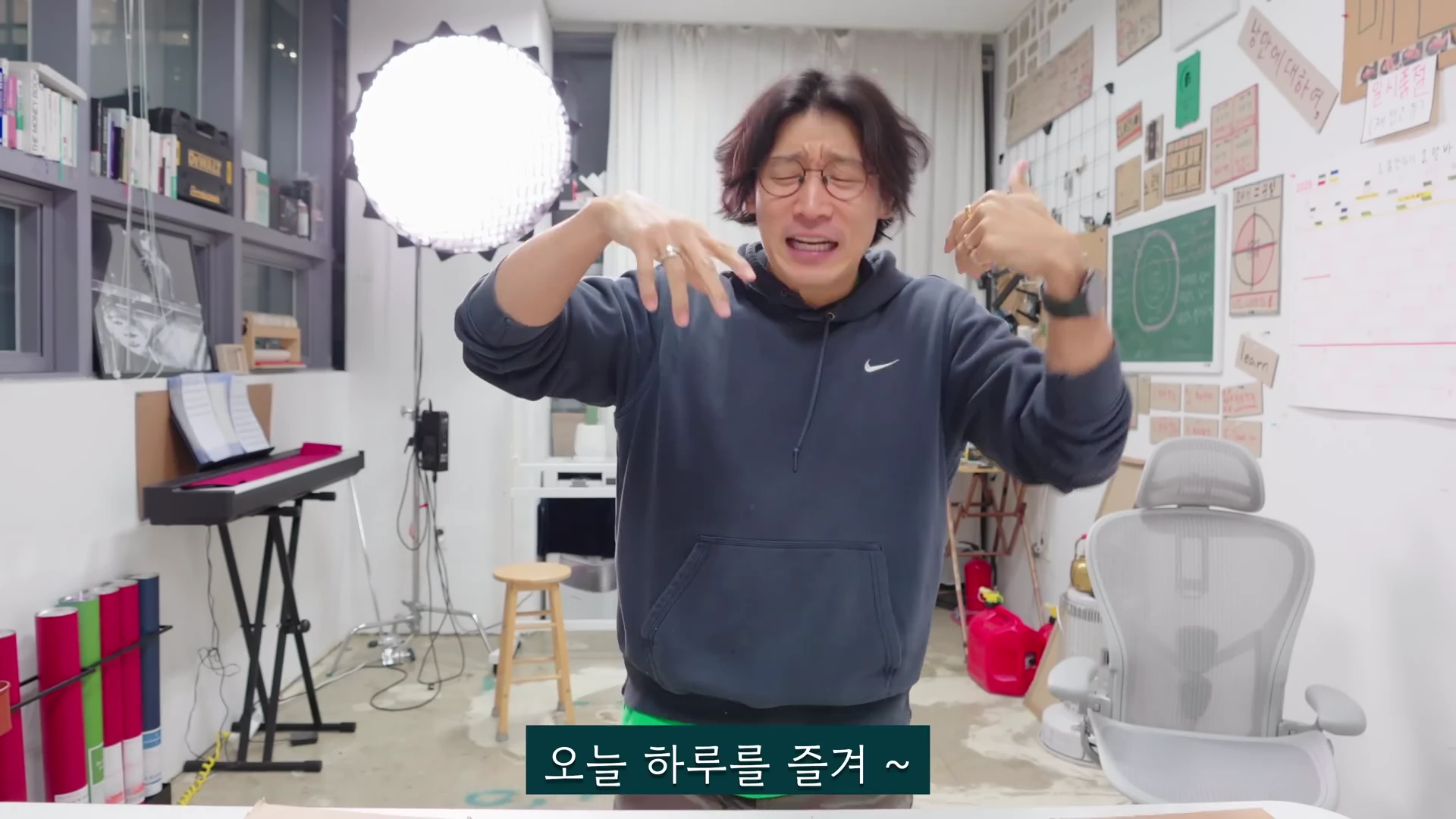 실내 스튜디오에서 어두운 후드티를 입은 남성이 안경을 쓰고 양손으로 원을 그리듯 제스처를 하며 이야기하고 있다. 배경에는 조명과 책장, 화이트보드가 놓여 있다.