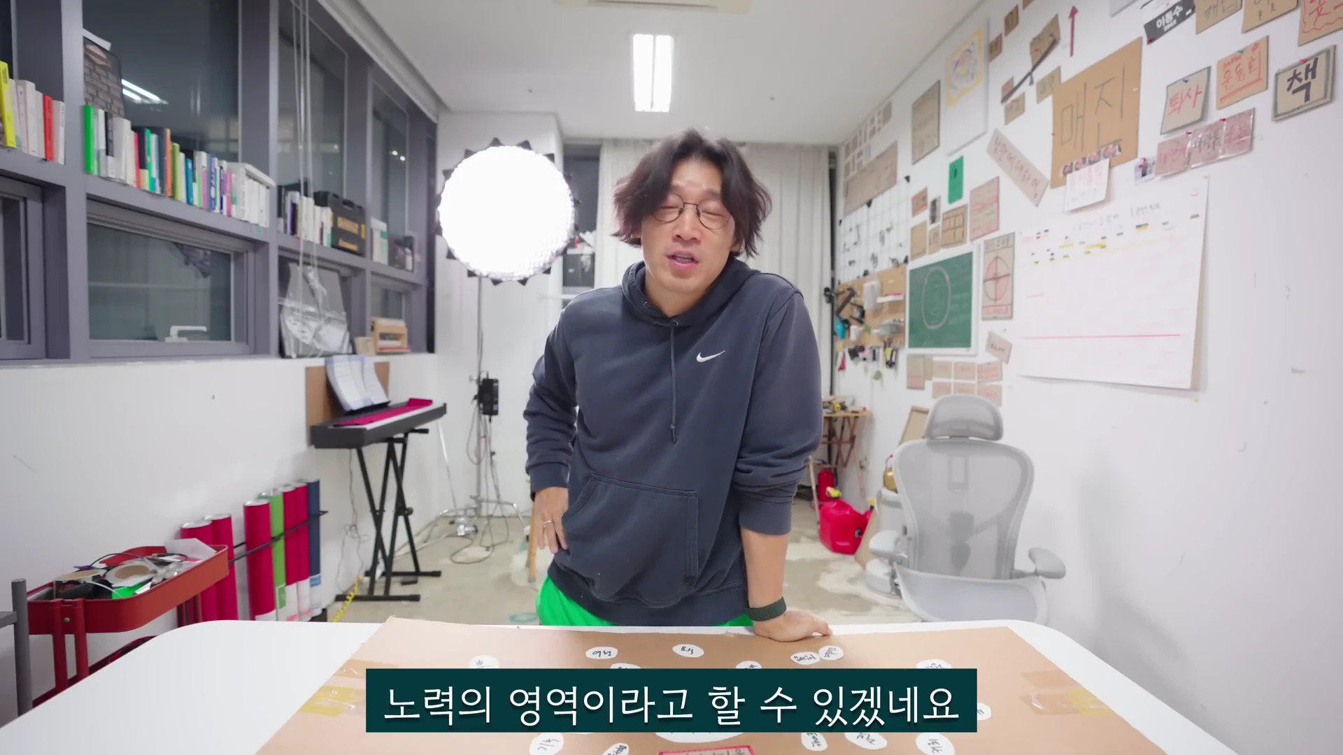 안경을 쓴 남성이 후드티를 입고 작업실에서 책상을 짚은 채 서서 이야기하고 있으며, 화면 하단에는 한국어 자막이 표시되어 있습니다.