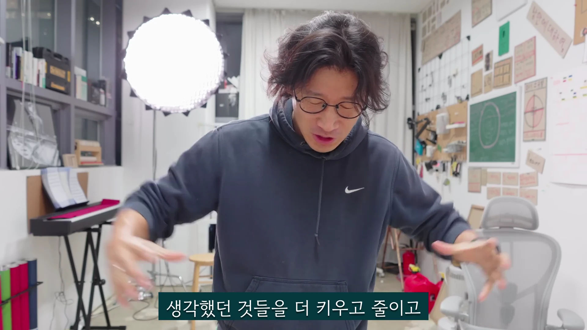 실내 작업실에서 후드티를 입은 안경 쓴 남성이 양손을 크게 벌려 무언가를 설명하는 모습이 담긴 영상 캡처 화면입니다.