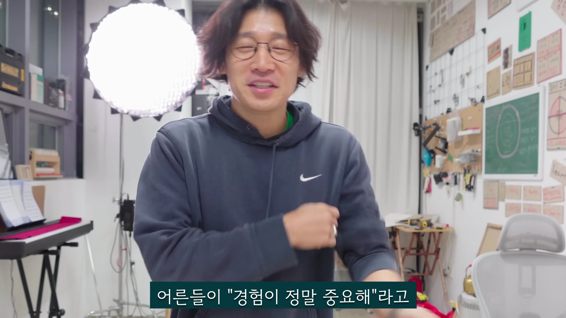 실내 스튜디오에서 편안한 후드티를 입은 안경 쓴 남성이 환하게 웃으며 손짓하며 이야기하고 있고, 하단에는 한국어 자막이 있는 영상의 한 장면입니다.