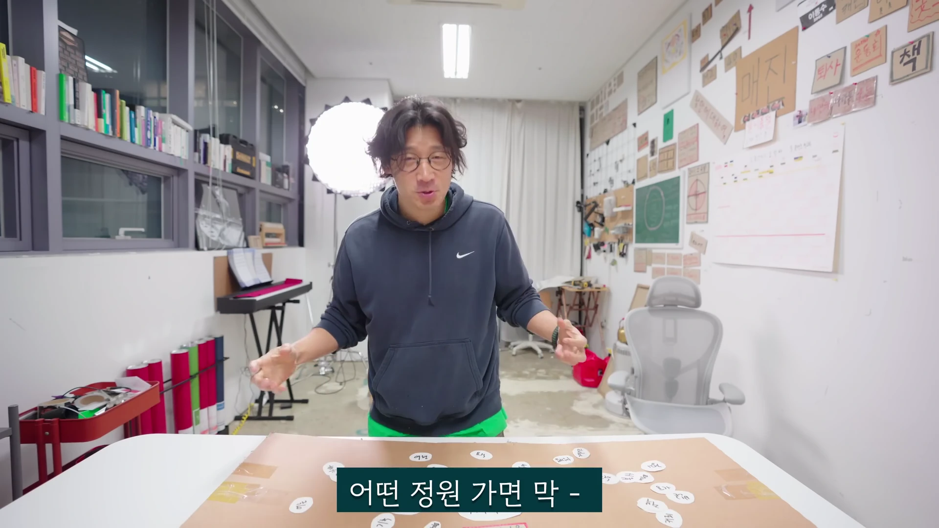 안경을 쓴 남성이 작업실 책상 앞에 서서 양손을 들어 설명하고 있으며, 책상 위에는 여러 개의 종이 조각이 놓여 있고 벽면에는 메모지가 가득 붙어 있습니다.