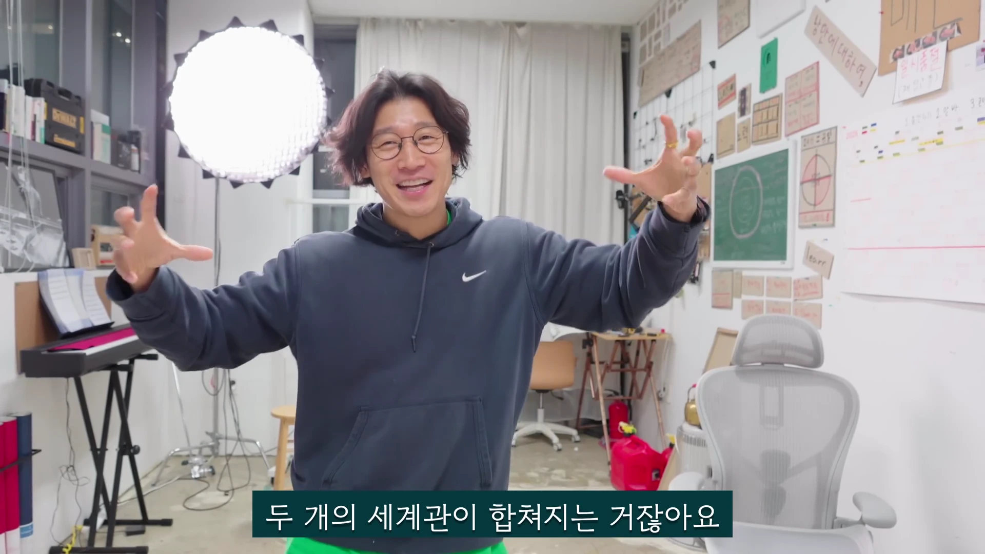 실내 작업실에서 안경을 쓰고 후드티를 입은 남성이 양손을 크게 벌려 설명하는 모습의 영상 캡처
