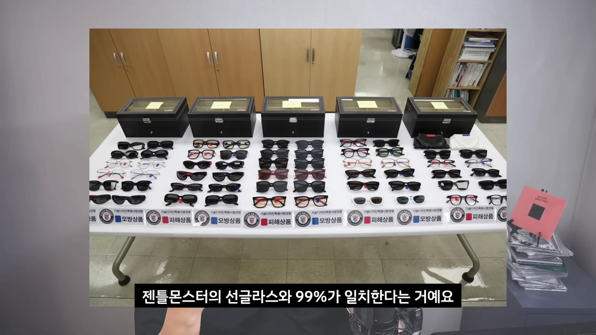흰색 테이블 위에 진열된 다수의 선글라스와 각 제품 아래에 '모방상품' 혹은 '피해상품'이라고 적힌 안내 문구가 놓여 있고 그 뒤로 검은색 안경 보관함들이 일렬로 배치된 모습.