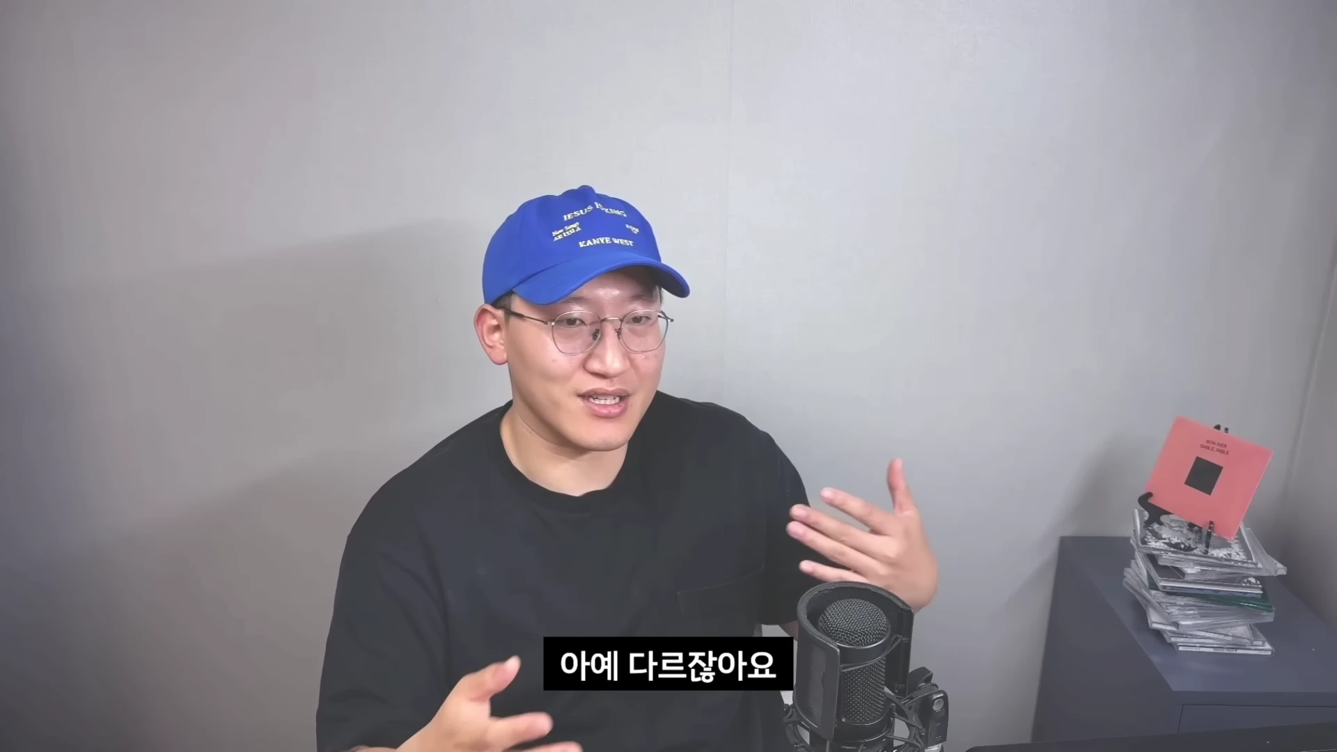 파란색 볼캡을 쓰고 검은색 티셔츠를 입은 남성이 마이크 앞에서 손짓하며 이야기하고 있는 모습