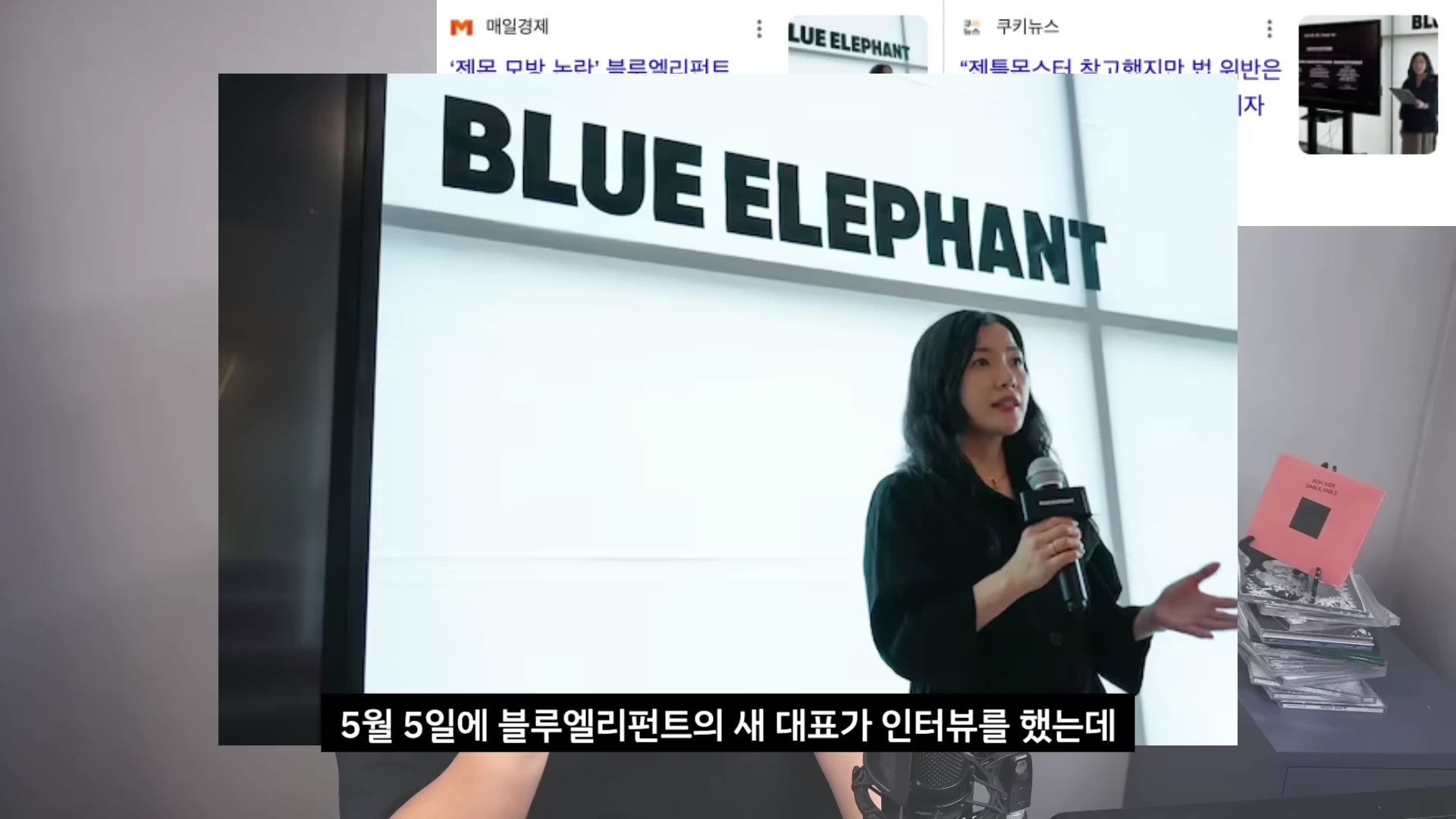 브랜드명이 적힌 큰 벽면을 배경으로 한 여성이 마이크를 들고 발표하는 모습이 담긴 화면