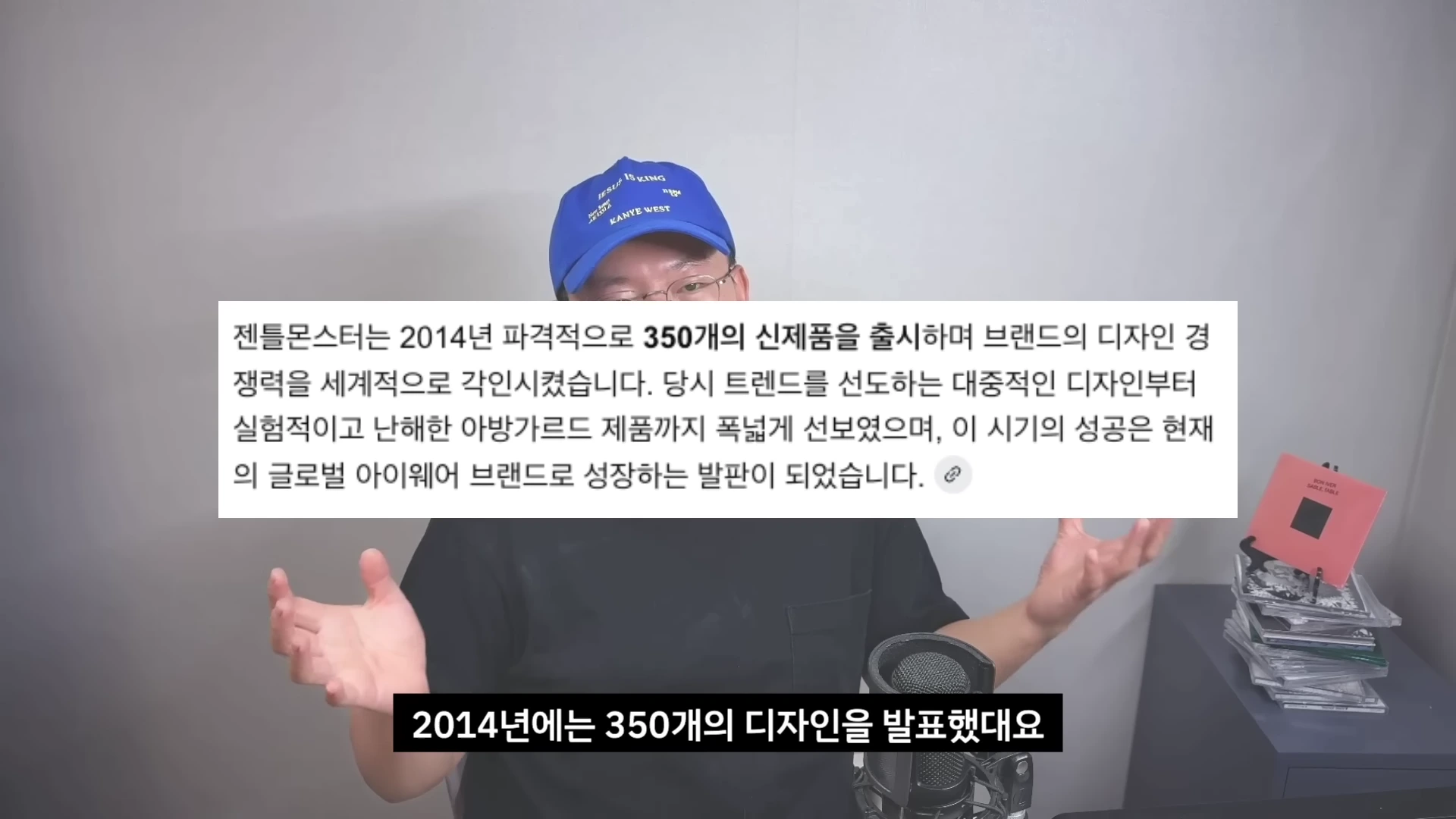 파란색 모자를 쓴 남성이 마이크 앞에 앉아 양손으로 제스처를 취하고 있으며, 화면 중앙에는 2014년 브랜드의 디자인 전략에 대한 텍스트가 삽입된 영상 캡처 화면입니다.