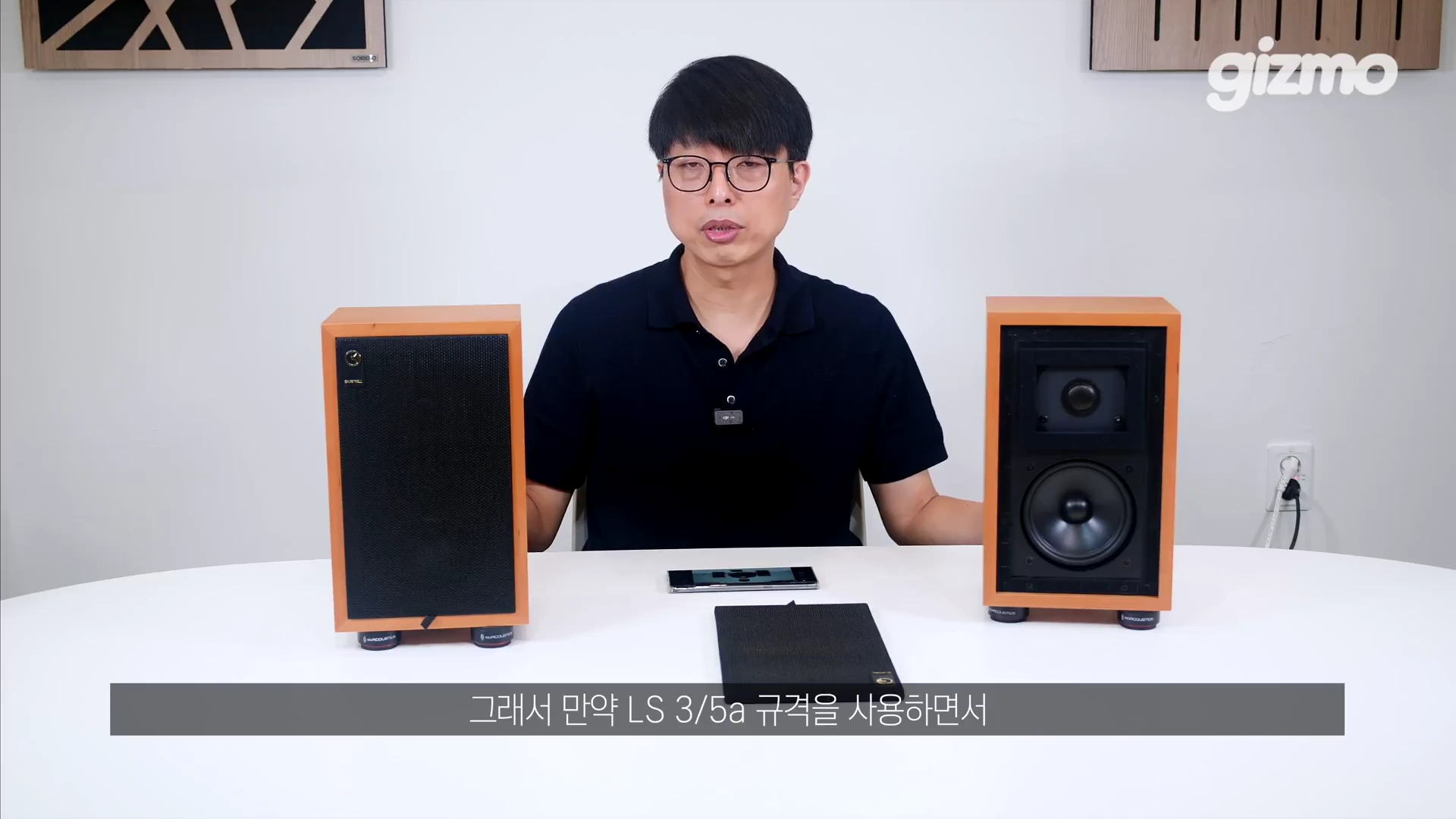 흰색 배경의 스튜디오에서 안경을 쓴 남성이 양옆에 목재 마감 스피커를 두고 설명하고 있으며, 한쪽 스피커는 그릴이 덮여 있고 다른 쪽은 유닛이 노출된 상태임