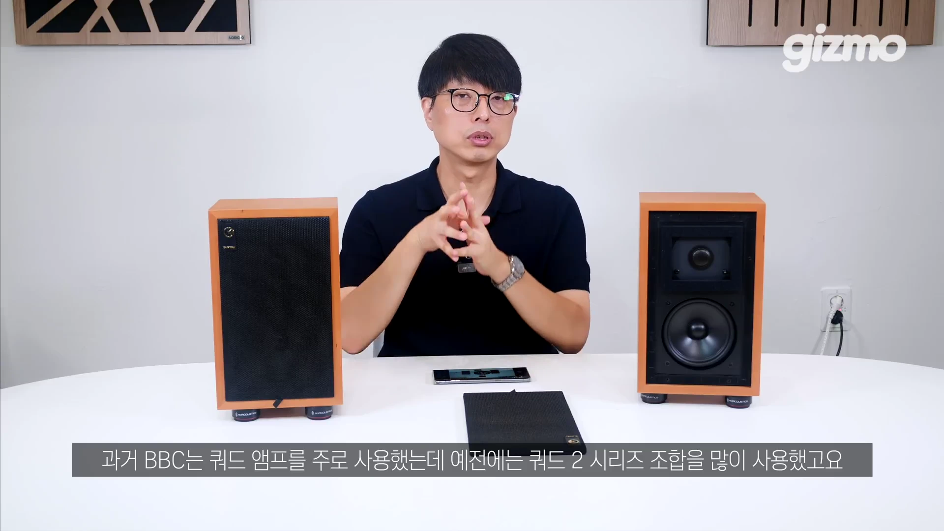 안경을 쓴 남성이 하얀 테이블 뒤에 앉아 있고, 양옆에 우드 마감의 스피커가 놓여 있으며 한쪽은 그릴이 제거된 상태로 촬영된 스튜디오 영상 화면입니다.