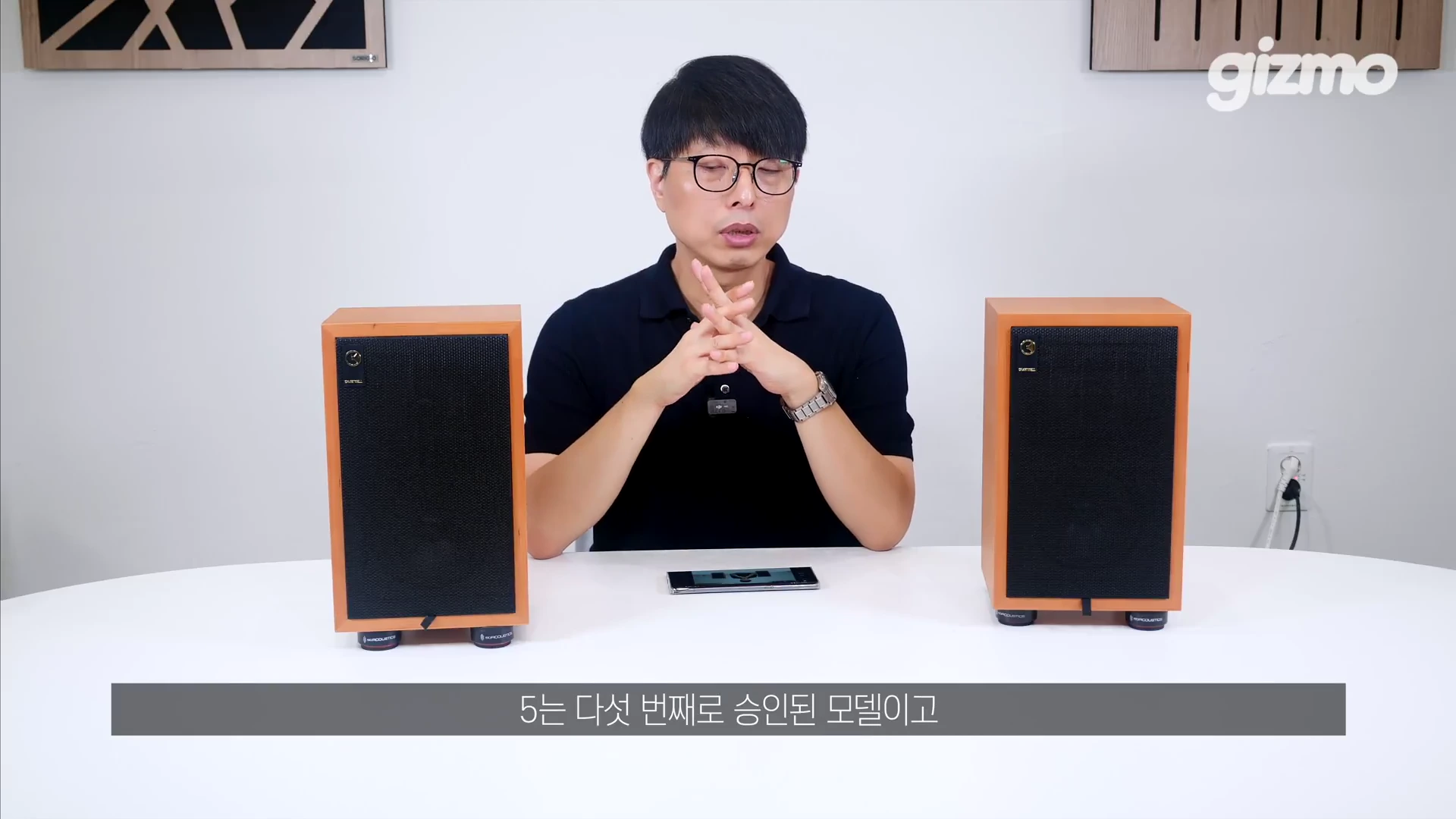 흰색 테이블 위에 그라함 차트웰 LS 3/5a 스피커 두 대가 놓여 있고, 그 뒤로 검은색 안경을 쓴 남성이 앉아 설명하고 있는 영상 캡처 화면입니다.