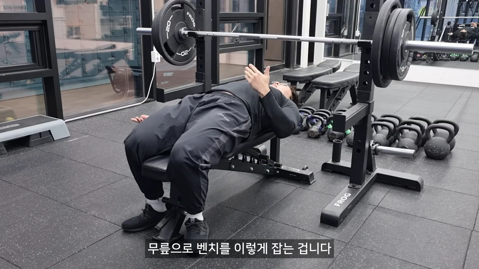 벤치프레스 랙 앞에 누운 남성이 양 무릎을 굽혀 벤치 양옆을 조이듯 고정하는 시범을 보이고 있다.