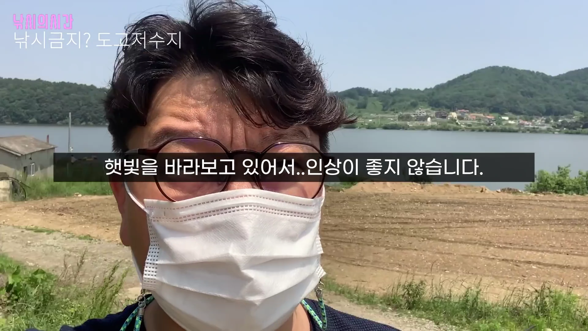 안경과 마스크를 쓴 남자가 저수지를 배경으로 셀프 촬영을 하고 있으며 화면 상단과 중앙에 텍스트가 삽입된 영상 프레임입니다.