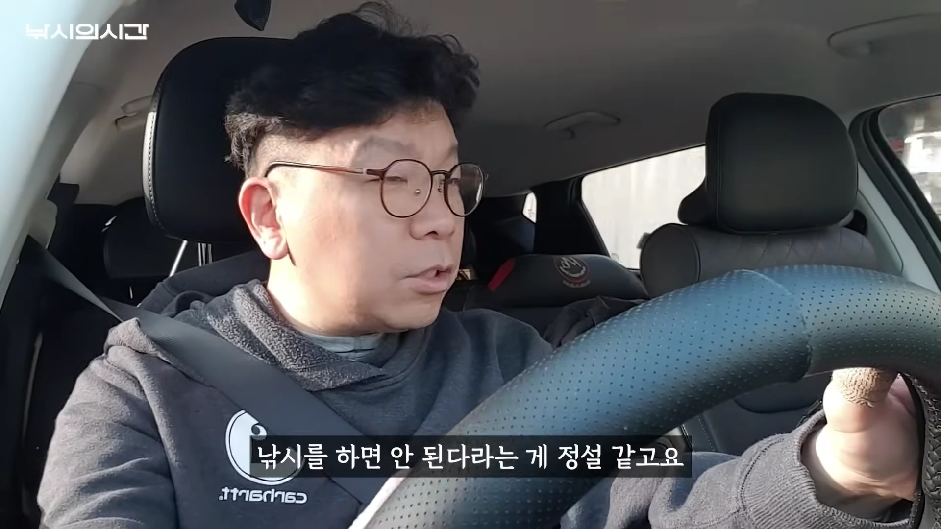 안경을 쓴 남성이 차 안에서 운전대를 잡고 이야기하고 있는 모습